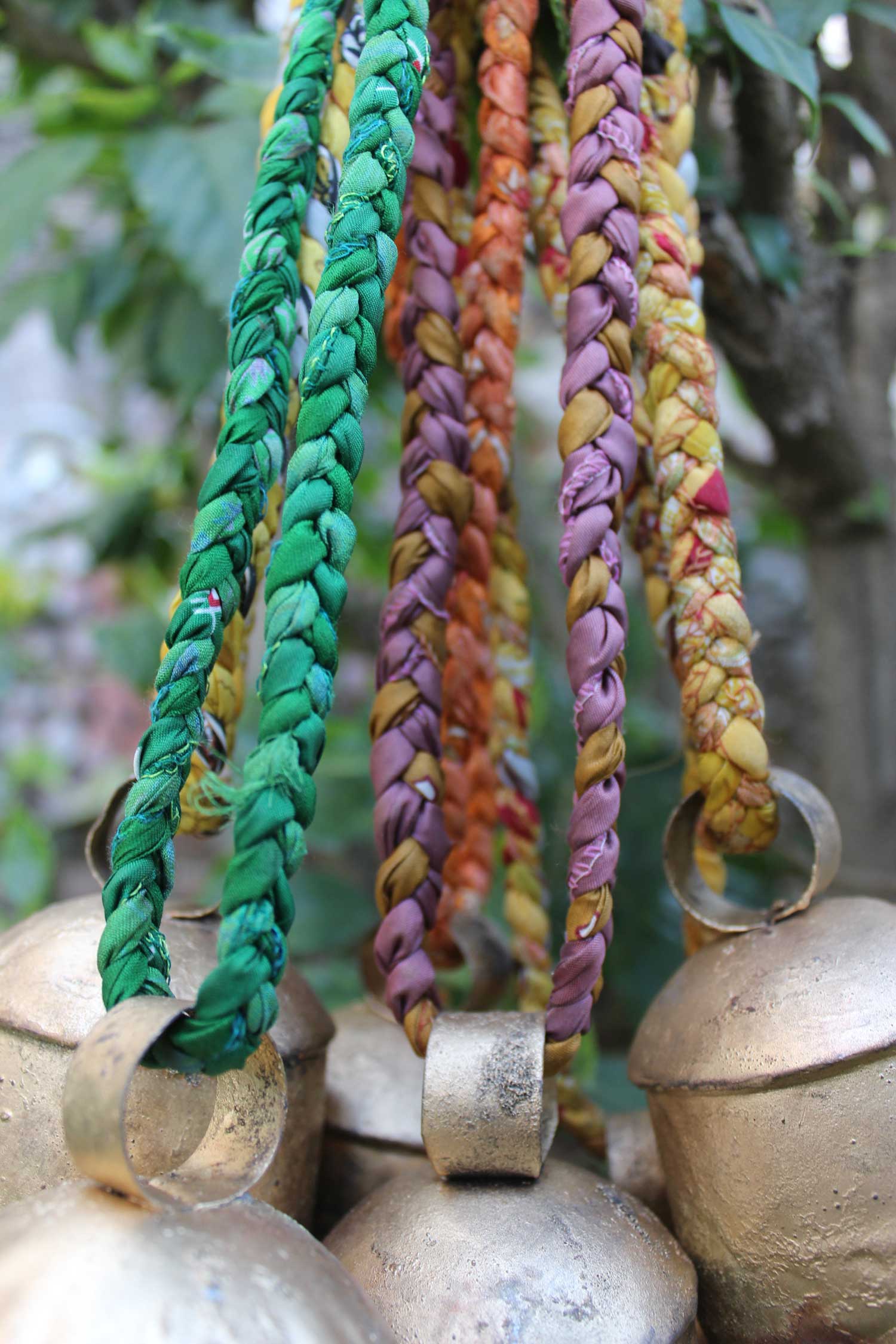 Colorful braided sari handles attached to rustic vintage tin cow bells, perfect for outdoor hanging decor.