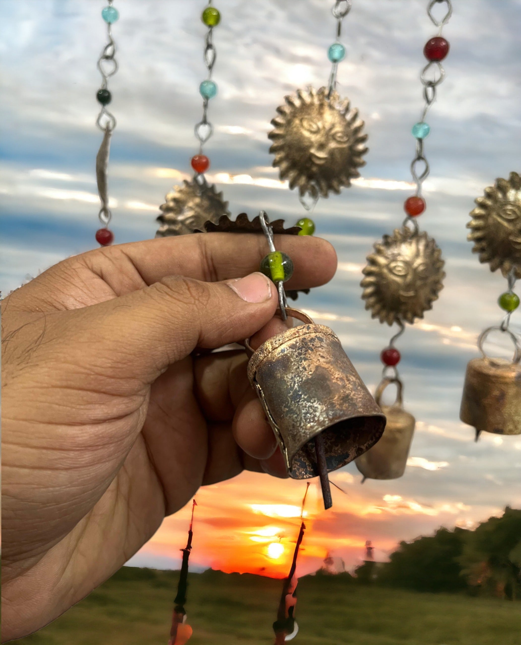 Hand holding a decorative metal wind chime with assorted bells and colorful beads, set against a sunset backdrop.