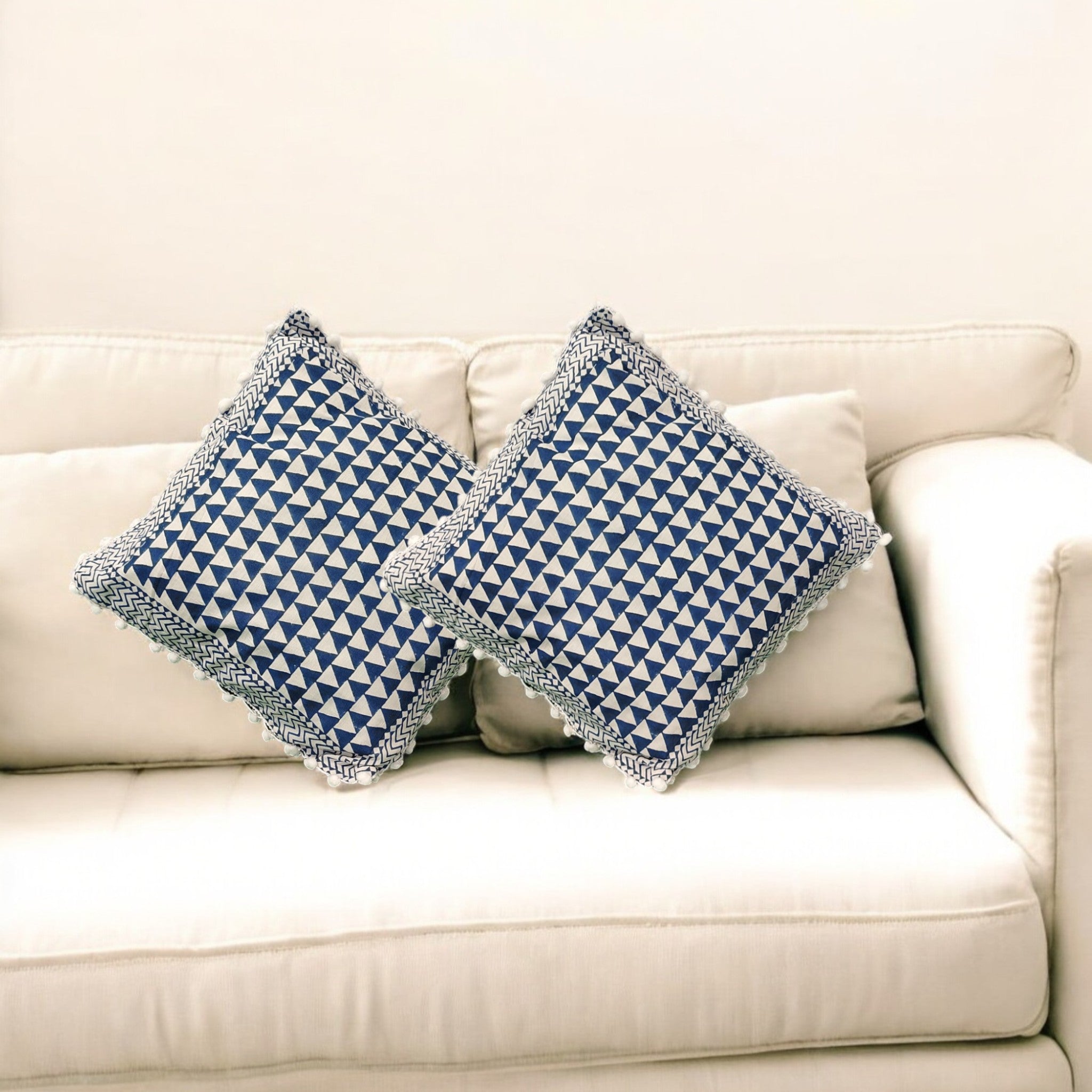 Set of 2 blue and white geometric triangle cushion covers with pom-pom fringe, styled on a beige couch.