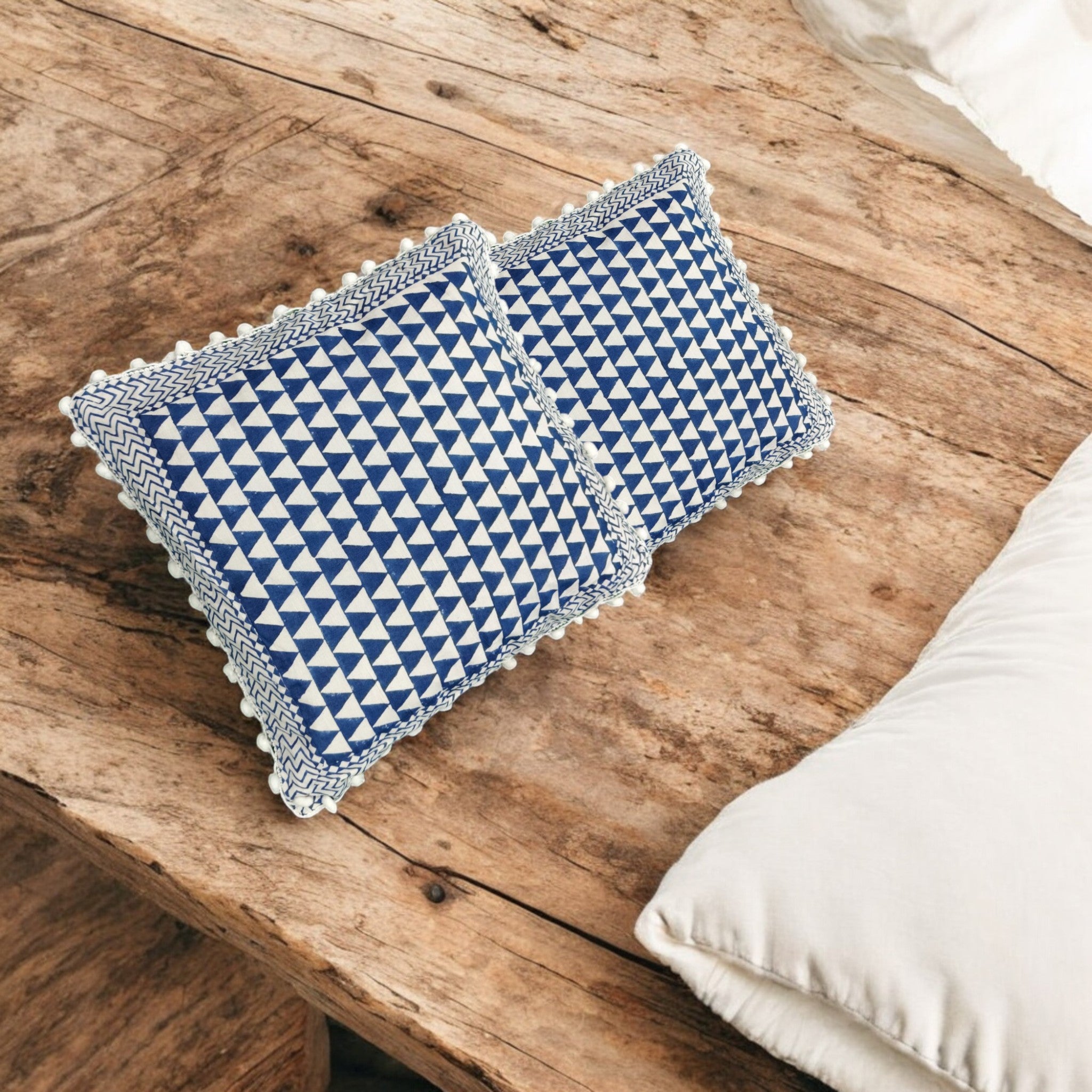 Set of 2 blue and white geometric printed cushion covers with pom-pom fringe on a wooden table.