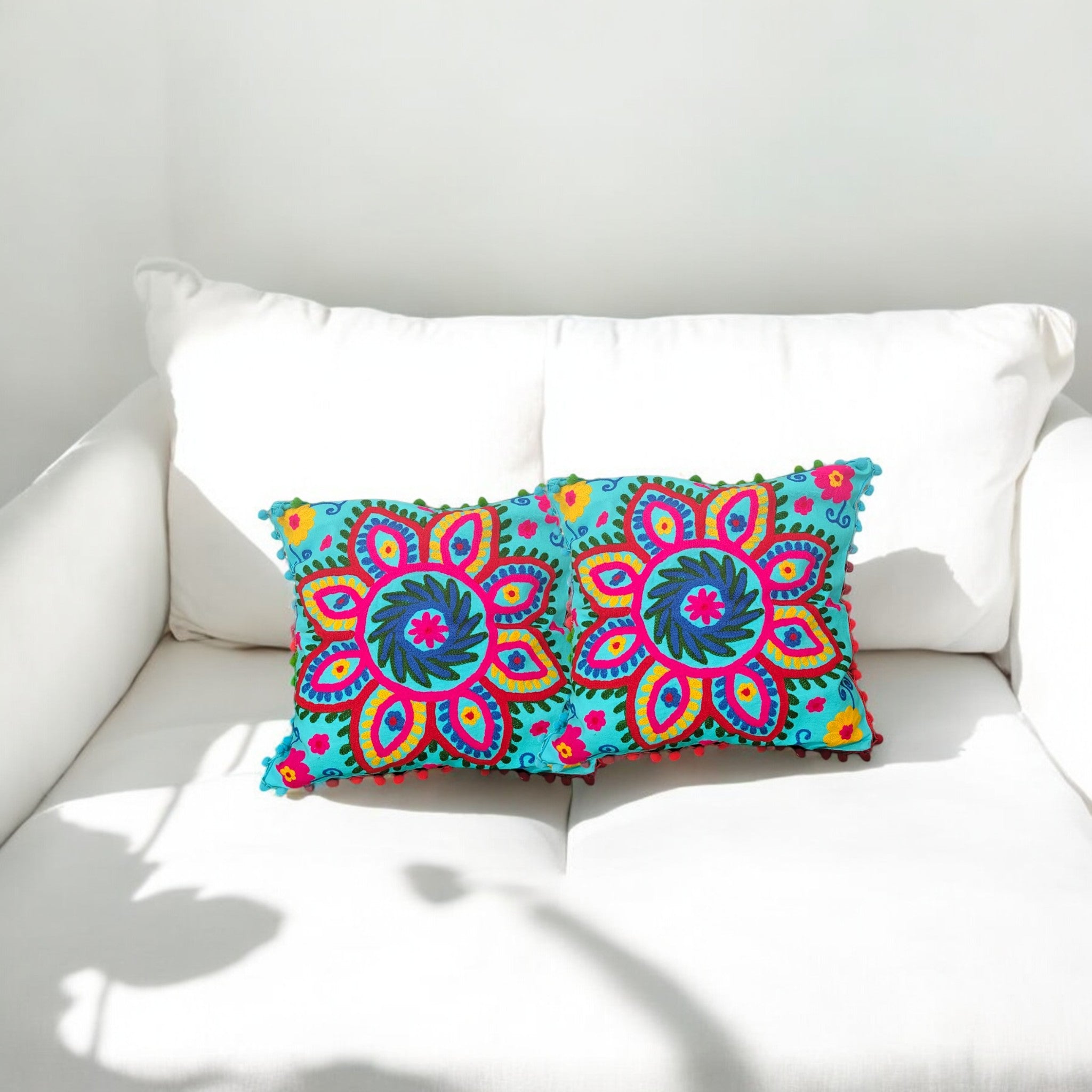 Set of 2 multicolor floral embroidered cushion covers on a white sofa, bright blue base with pom-pom edges.