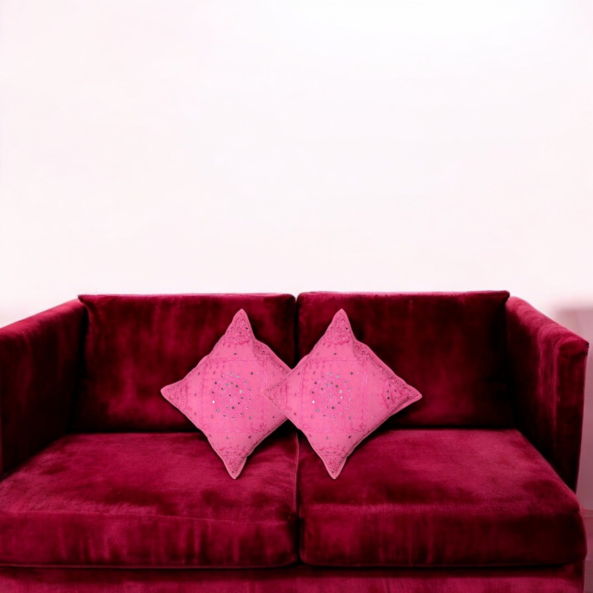 Set of 2 pink mirror work cushion covers on a plush burgundy sofa, adding a pop of color to home decor.
