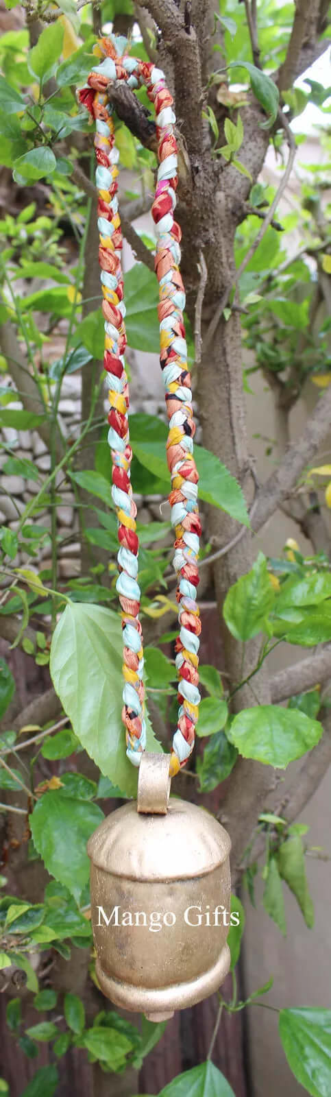 Handcrafted recycled iron bell hanging from a decorative braided cord against green foliage.