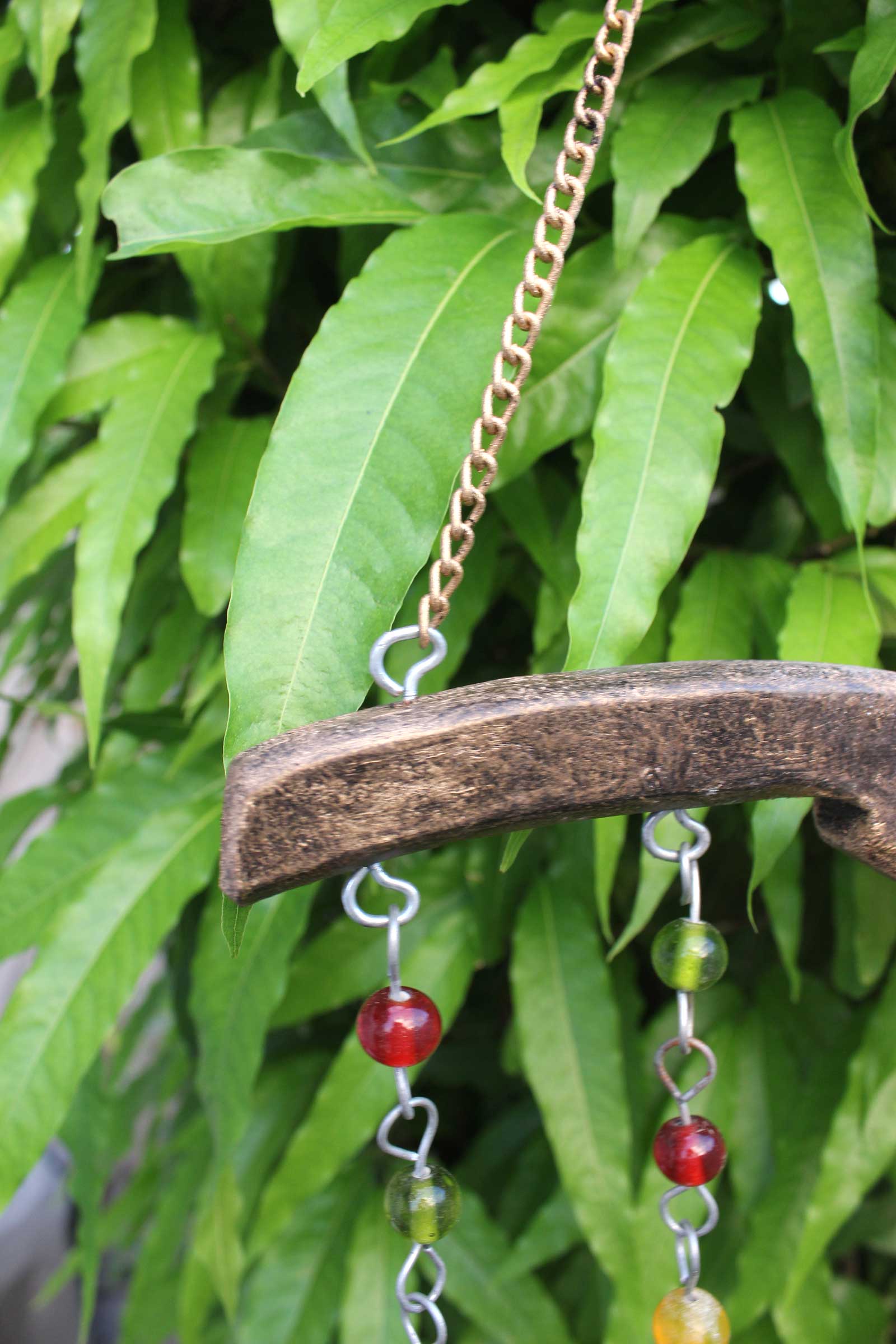 Handmade beaded wind chime hanging from wood, featuring colorful glass beads and a chain, surrounded by lush green leaves.