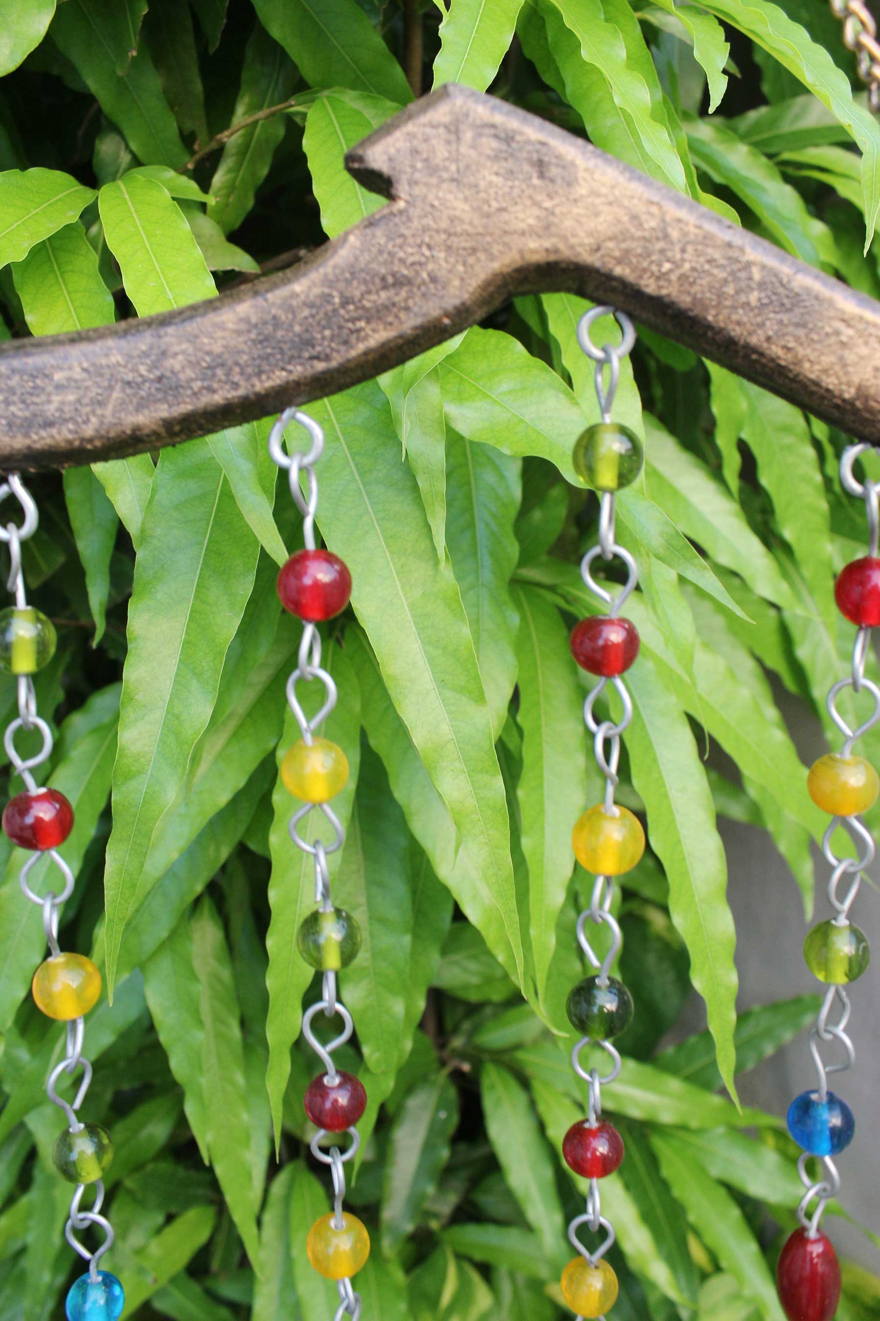Colorful glass beads on a handmade hanging chime with wooden hook, set against green foliage.